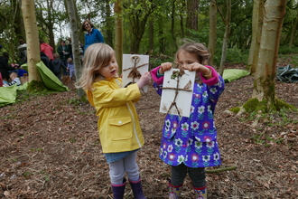 2 girls hold up nature craft painting they made in forest other people in background