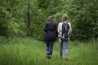 Two women walking away from the camera into woodland, one has a rucksack on, they look deep in conversation.