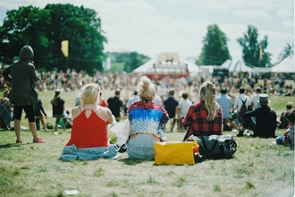 An outdoor summer festival with people sat on the grass in the sun