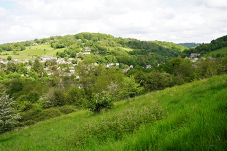 The view from an area of grassland, towards a mosaic of trees, houses, and another hill in the background.