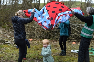 In a nature reserve. Spotty ladybird parachute being lifted by adults. Child smiling and running under the parachute.