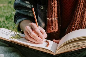 Someone writing in a book, they are wearing a scarf and sat outside