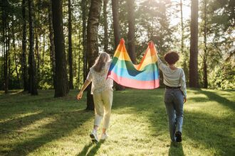 Two people walk through a forest holding a rainbow flag between them.