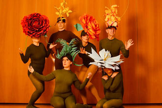 A group of 6 people in dark green leotards with large flower head pieces