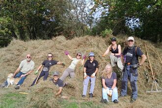 A group of people sat and standing in front of some cut hay and trees in the background