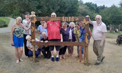 a group of people stood around a photo frame with "mission wild" written at the top, the sign is made out of wood and there is grass and trees in the background