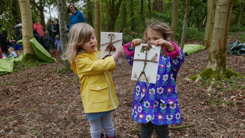 2 girls hold up nature craft painting they made in forest other people in background