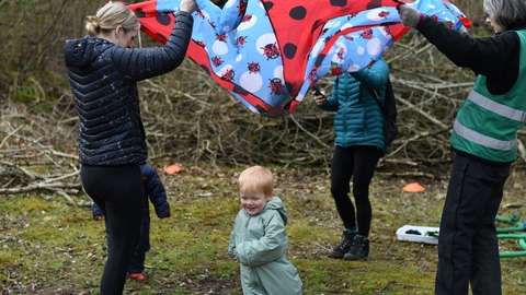 In a nature reserve. Spotty ladybird parachute being lifted by adults. Child smiling and running under the parachute.