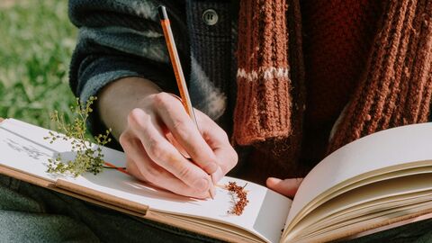 Someone writing in a book, they are wearing a scarf and sat outside