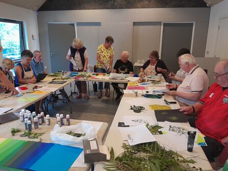 A group of people sat round a U shaped table with pieces of paper, paint and leaves in front of them