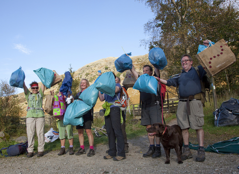Litter picking organized event multiple volunteers holding up blue bags with litter/rubbish inside. Accompanied by one brown Labrador dog. A hill is shown in the backdrop along with a few trees. 