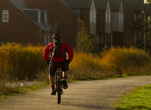 Man cycling down path by a river with houses in background