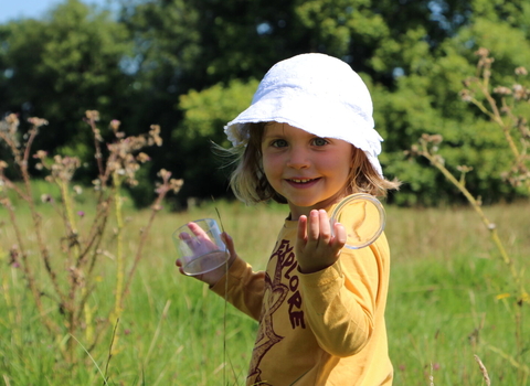 Girl_with_bug_pot in tall grass wearing white hat looking into camera